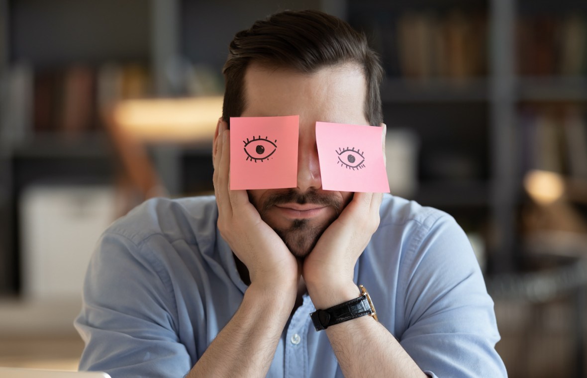 Bărbat doarme la birou cu două post-it-uri pe care sunt desenați ochi deschiși