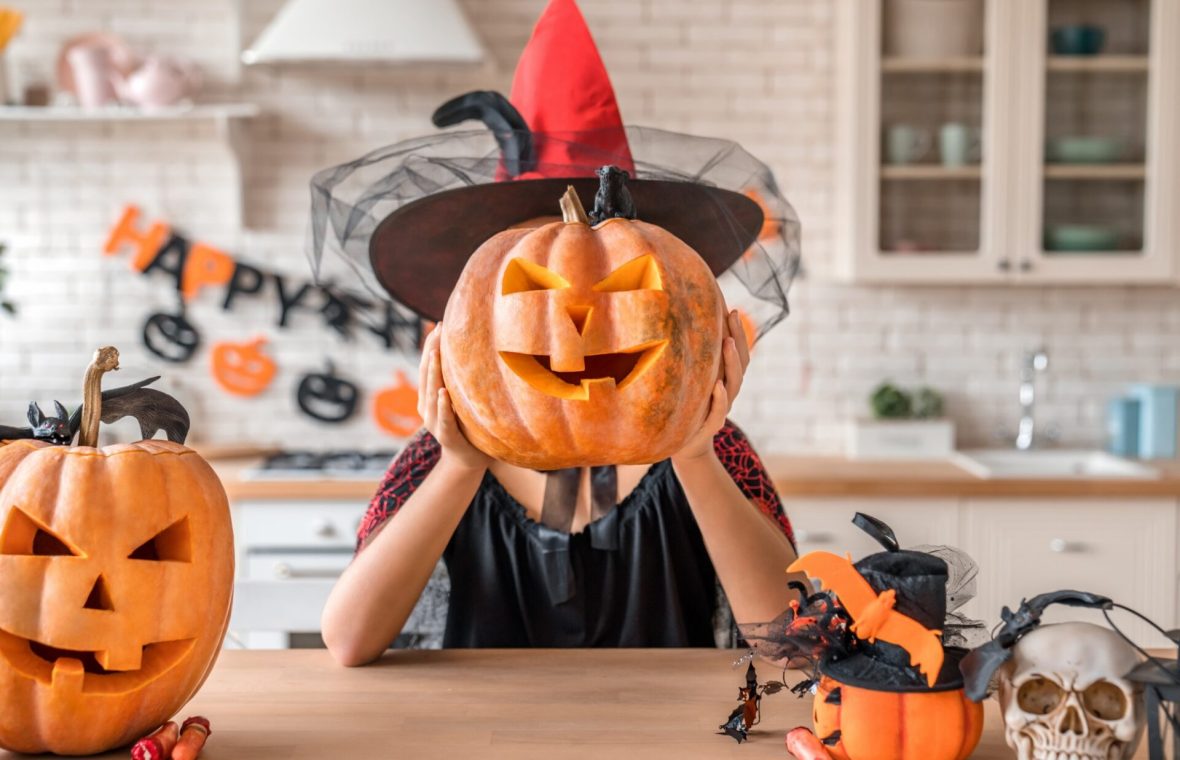 dovleci de halloween sculptați, femeie costumată în vrăjitoare ține un dovleac sculptat în dreptul capului