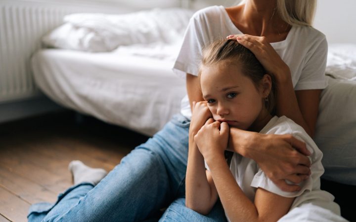 Mama îmbrățișează strâns fiica tristă, stând pe jos, lângă pat