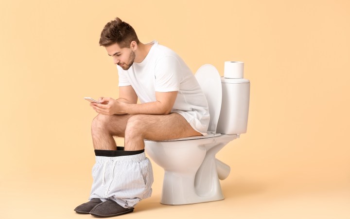 bărbat stă pe vasul de toaletă cu telefonul în mână