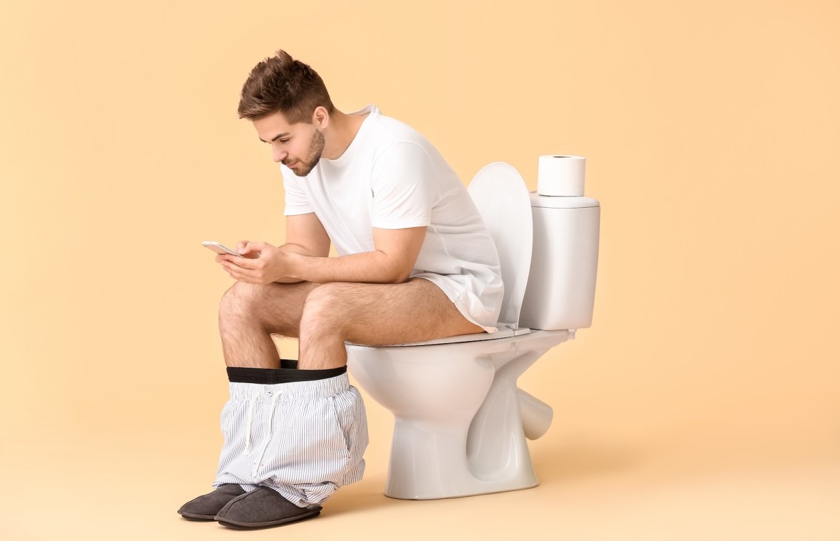 bărbat stă pe vasul de toaletă cu telefonul în mână