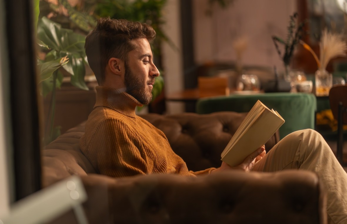 bărbat într-un pulover pe gât citind o carte pe canapea într-o cafenea cozy, poză din profil