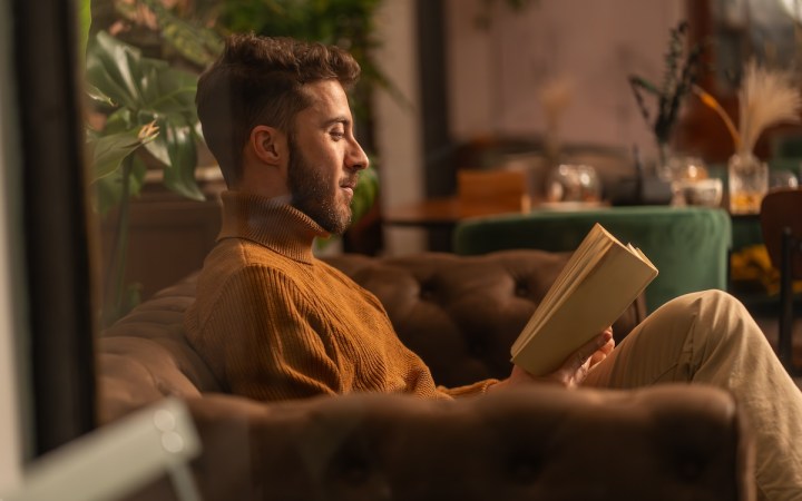 bărbat într-un pulover pe gât citind o carte pe canapea într-o cafenea cozy, poză din profil