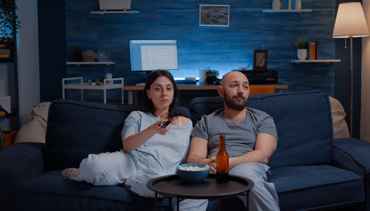 Parteneri de cuplu îmbrăcați în pijamale plictisiți, mănâncă popcorn, el bea bere, ea schimbă canalele la televizor, el se uită într-o parte