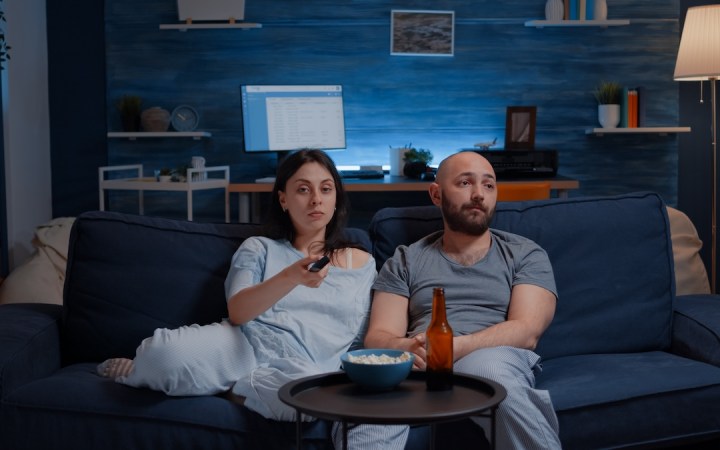 Parteneri de cuplu îmbrăcați în pijamale plictisiți, mănâncă popcorn, el bea bere, ea schimbă canalele la televizor, el se uită într-o parte
