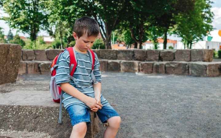 Copil trist, singur pe stradă, roșu în obraji și cu ghiozdanul în spate