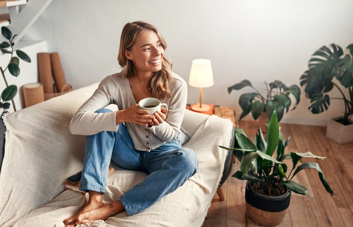 ce înseamnă o zi fericită? femeie care zâmbește în timp ce bea o cafea acasă și e înconjurată de lumină și plante