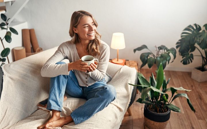 ce înseamnă o zi fericită? femeie care zâmbește în timp ce bea o cafea acasă și e înconjurată de lumină și plante
