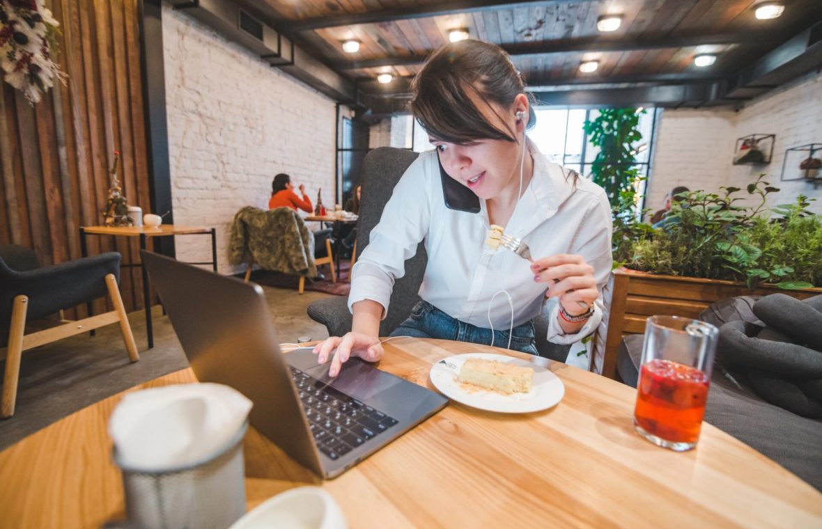 femeie în ședință, la laptop, cu telefonul la ureche, ia prânzul la cafenea