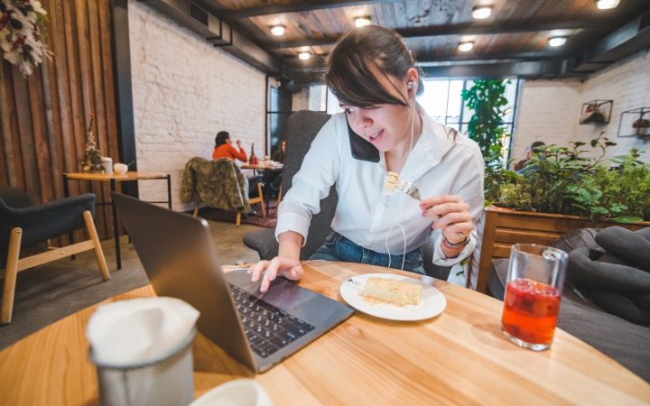femeie în ședință, la laptop, cu telefonul la ureche, ia prânzul la cafenea