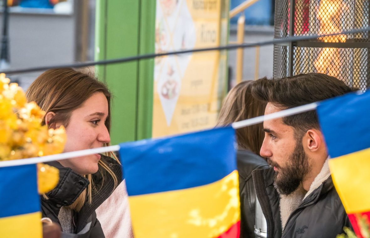 Un cuplu discută într-un cadru public decorat cu steaguri tricolore, ilustrând tensiunile și conversațiile tot mai frecvente legate de politica în relații, mai ales în contextul alegerilor din România