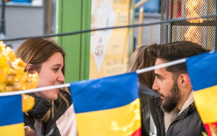 Un cuplu discută într-un cadru public decorat cu steaguri tricolore, ilustrând tensiunile și conversațiile tot mai frecvente legate de politica în relații, mai ales în contextul alegerilor din România