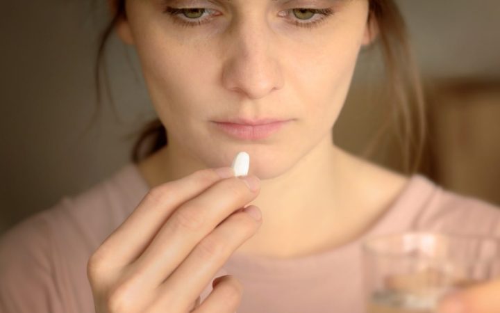 femeie foarte obosită, cu cearcăne care ia un medicament alb și pare că se uită în ecranul unui calculator