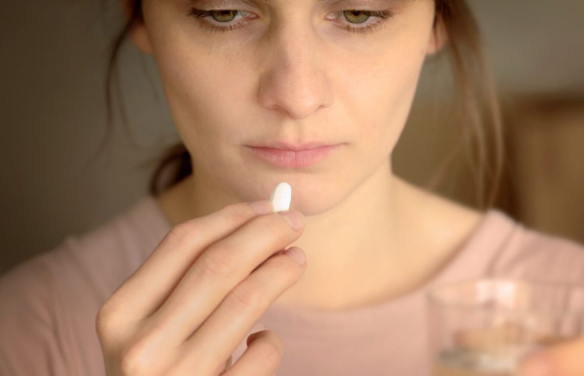 femeie foarte obosită, cu cearcăne care ia un medicament alb și pare că se uită în ecranul unui calculator