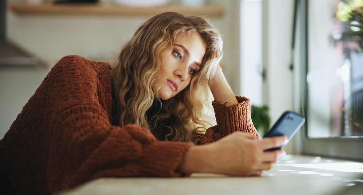 femeie tânără tristă se uită pe telefon în casa ei singură