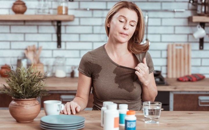femeie blondă cu pete de transpirație pe tricou un pahar cu apă pe masă li cutii cu pastile