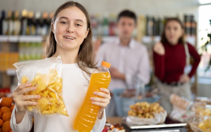 adolescentă care ține într-o mână o pungă cu chipsuri și în cealaltă un suc portocaliu