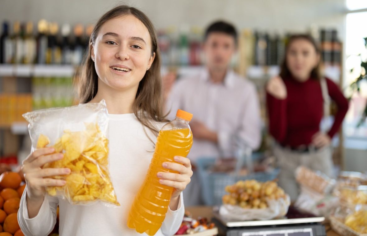 adolescentă care ține într-o mână o pungă cu chipsuri și în cealaltă un suc portocaliu