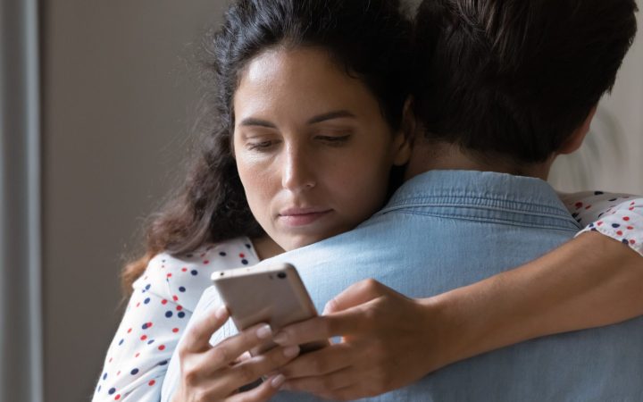 cât de monogami sunt oamenii? femeie care își verifică telefonul în timp ce-și îmbrățișează partenerul