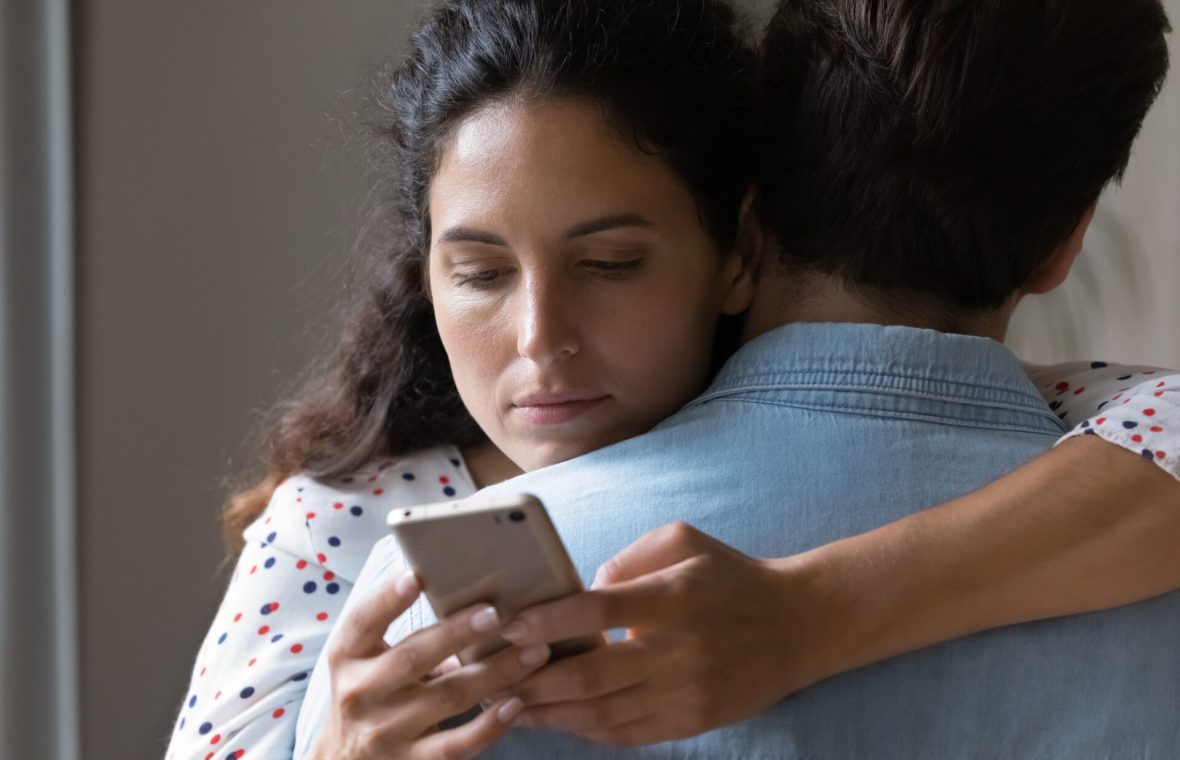 cât de monogami sunt oamenii? femeie care își verifică telefonul în timp ce-și îmbrățișează partenerul